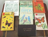 Seven vintage books arranged on a table, covers visible, titles related to herbs, birds, marijuana, trees, insects, butterflies, and wild flowers.