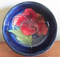 Top-down view showing red Clematis flower with green leaves on cobalt blue background inside bowl.