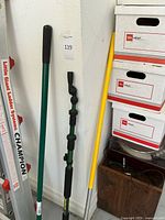 Photo showing shovel and green garden rake leaning against wall next to stacked boxes.