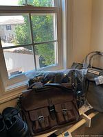 Three bags and pair of black slip-on shoes on window bench: grey canvas tote with Barnes & Noble label, brown leather satchel, black shoes, and protective dust bag with shoebox.