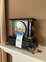 Front view of black decorative clock with gold/brass accents and round clock face with Roman numerals, visible label with size noted as 10x12x6 inches, and winding key on shelf.