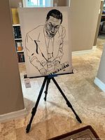 Front view of black metal artist easel with adjustable legs holding a signed monochrome canvas portrait of a man playing piano.