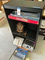 Full view of black bookcase with board games on upper shelf, cuckoo clock in middle shelf, and electronics on bottom shelf