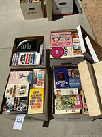 Four open cardboard boxes filled with paperback books placed on a concrete surface. Visible titles and authors span various genres.
