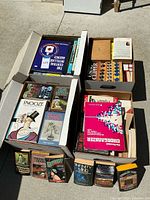 Four open boxes shown from above with various books arranged inside on pavement. Includes titles in espionage, military, reference, and programming categories.