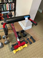 Exercise bench with 8 dumbbells placed around it, showing black and yellow coated dumbbells and the red frame of the bench.