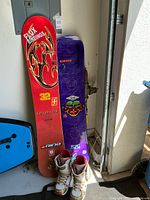 Two snowboards and a pair of snowboard boots positioned upright on the floor against a wall, showing logos and graphics for Joyride and Lib Tech, and white Burton boots in front.