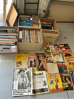 Four cardboard boxes filled with magazines, paperbacks and hardcovers plus laid-out issues on floor