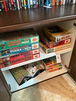 Photo shows multiple puzzle boxes stacked on two shelves inside a wooden cabinet. Puzzle boxes include Interlaken Super Large Puzzle 2500 pieces, Super-Luxe Puzzle 2500 pieces, and others labeled 1500 and 1000 pieces. Also visible are Big Ben board games in worn boxes and York boxes likely for models or kits.