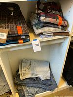 Top shelves show stacked men's button-down shirts in a variety of colors and patterns including plaid, striped, flannel, and solid. Many have size labels showing XL, XXL and Large. Some shirts have tags indicating they are new.