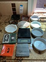 Lot of mixed ashtrays, dishes, shell, wood trays and scale arranged on a countertop under window light.