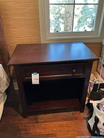 Full view of dark brown wooden nightstand under a window, showing front with single drawer and open shelf below, tags visible on drawer and shelf.