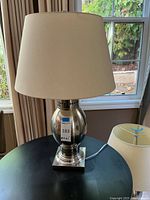Full view of one table lamp showing silver metal base and off-white fabric shade on a black surface near a window.