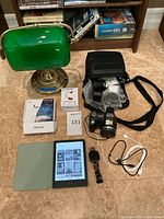 Sony digital camera with lens, carrying case, Amazon Kindle e-reader in cover, Fitbit Versa smart watch, charging cables, and green desk lamp on floor