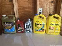Five containers of automotive fluids including antifreeze/coolant in large yellow Prestone jugs, motor oil in various brand bottles, and synthetic oil