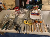 Overall view of assorted painting supplies on table, showing paint trays, rollers, brushes, putty tool, Paint Paul, rags, and tape.