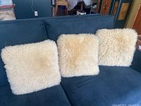 Three Auskin sheepskin pillows arranged on a dark blue couch, showing their cream-colored soft fur surfaces with texture variations.