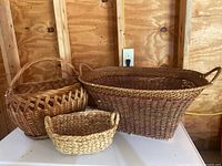 Front and side view showing three baskets of different sizes arranged on a white surface against a wooden wall. The largest basket shows its oval shape and handle condition.