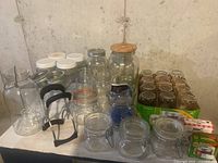 Glass canning jars of various sizes and types on a table, including wire clamp jars, white plastic lid jars, and metal lid jars. Also visible are black jar clips and few extra lids.