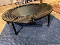 Mini-trampoline rebounder with black padded edge and foldable legs on a blue carpeted floor