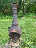 Full view of rusty brown metal Chimera outdoor fireplace standing on concrete blocks in a grassy outdoor setting.