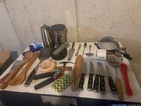 Overview of assorted kitchen utensils, knives, and cooking tools laid out on a table, showing a variety of wooden, metal, and plastic items.