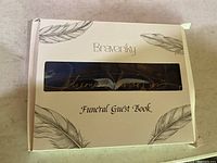 Front view of the Braveriky Funeral Guest Book in its box showing feather designs and brand name with visible cursive script inside through clear window.