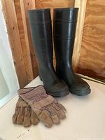 Photo of black rubber boots size 10 with slightly worn soles, standing upright on a surface with brown walls in background. Pair of work gloves with striped fabric backs and leather palms placed in front of boots.