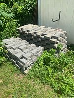 Two stacks of gray interlocking concrete garden pavers placed on the ground outside against a shed, surrounded by green plants.