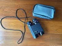 Photo showing black textured Konica binoculars with attached neck strap and gray zippered soft carrying case on wooden surface.