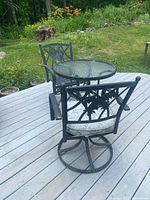 Outdoor round glass dining table with two black metal swivel chairs on a wood deck with garden in background