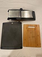 Photo shows OXO mandolin slicer with stainless steel blade and black plastic frame, alongside black plastic and bamboo cutting boards.