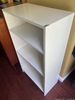 White particle board shelf unit showing top surface and adjustable holes on sides for shelves. Visible edge wear on top corner.