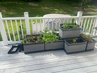 Wide view showing 8 of the rectangular gray plastic planters on a deck with plants inside. Some planters are stacked or paired side by side. Black stands visible nearby.