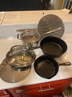 Overview shot of all items: All-Clad saucepan with lid, mesh splatter guard, three additional lids, two cast iron skillets (5 inch and 8 inch)