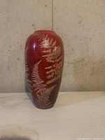 Front view of red ceramic vase with silver fern imprints on a light surface against a concrete wall background.