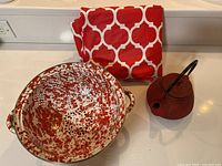 Photo showing all three items: red and white speckled metal strainer, red cast iron tea kettle, and folded red and white geometric patterned table cloth.