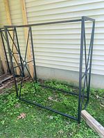 Photo showing one of two black metal firewood stackers against a house exterior on grass. Shows the rectangular frame and decorative metalwork sides.
