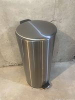 Full view of the round stainless steel garbage can with step pedal, against a concrete wall background.