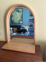 Front view of arch-shaped wooden mirror with bottom shelf, reflecting a room interior.
