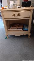 Front view of one cream-colored vintage French Provincial nightstand showing drawer with ornate handle and open shelf below containing books.
