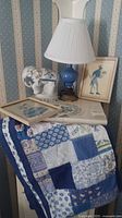 Blue ceramic lamp on a round wooden table with a white pleated shade, beside a white ceramic elephant figurine and two framed pieces of human figure art, the table runner underneath showing patchwork textile.