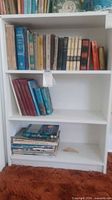 Photo showing full white painted bookshelf with three shelves, holding various books on the top and bottom shelves.