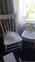 Photo showing two white wooden chairs, one with a damaged cane seat and the other with a solid seat with cushion, placed near a window and curtain.