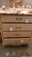 Front view of small 4-drawer chest with cream body and gold trim. Decorative gold handles and curved legs visible.
