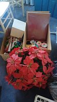 Photo showing a large artificial poinsettia wreath alongside open boxes containing Christmas bows, rolls of ribbon, and some gift boxes.