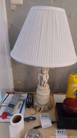 Full view of the cream cherub lamp on a cluttered surface showing the pleated lampshade and upper part of the lamp.