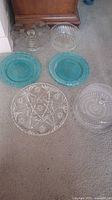 Top-down view showing five trays and cake stands including two turquoise blue glass plates and three clear glass/crystal trays with decorative patterns on carpet