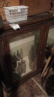 Display cabinet showing wood frame and glass doors with several decorative glass bottles inside, top surface with some objects including a white plastic basket.