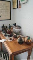 Wooden and ceramic duck figurines displayed on a wooden table, showing various sizes, realistic paint, and carving details.